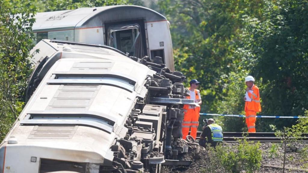 A train carriage on its side, with emergency workers stood next to it and a police officer crouching down next to it