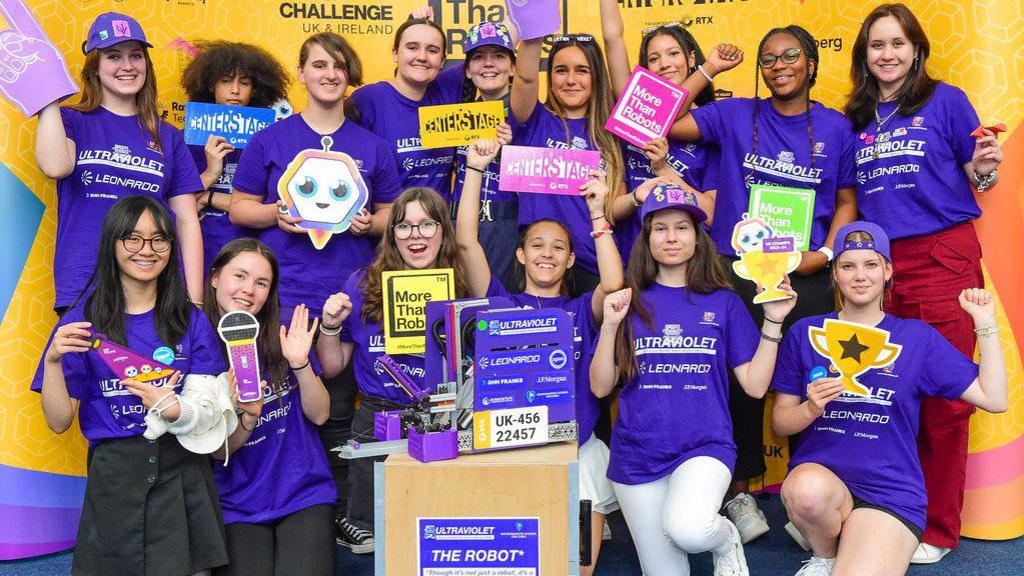 Fifteen teenage girls posing for the camera with a square-shaped robot. They are all wearing violet-colour t-shirts printed with the word Ultraviolet - the name of their team