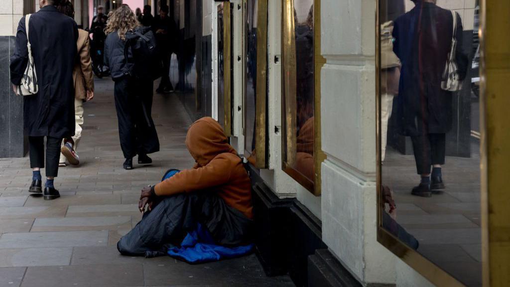 Homeless person seen sitting on the ground along a London street. He is wearing a yellow hoodie and looking away from the camera keeping his face concealed. Pedestrians can be seen walking along the street.