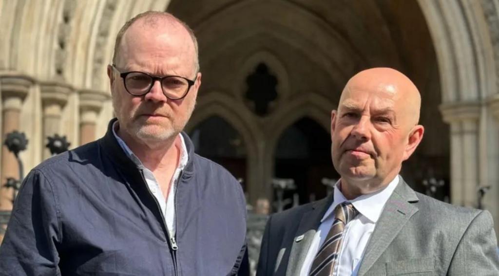 Two men face the camera. One is wearing a dark jacket and white shirt. He has dark rimmed glasses and a grey short beard. The man on the right is wearing a grey suit, white shirt and grey tie. He is balding.