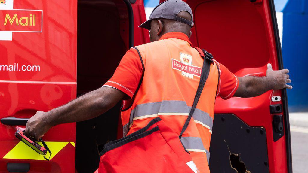 Postal worker wearing orange hi-viz with Royal Mail logo on the back shuts a postal van door   