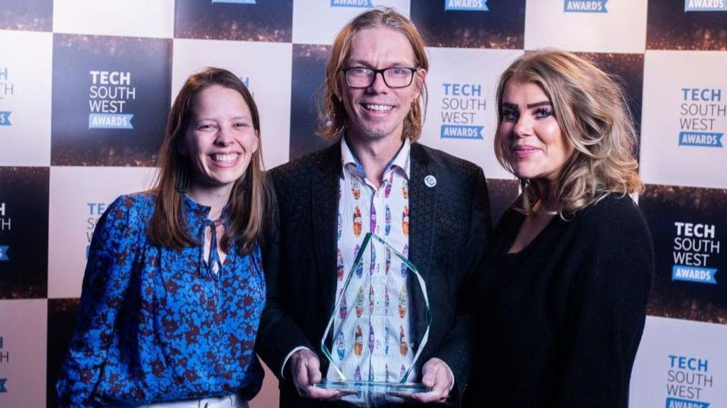 A group of three people from Bristol company Neurosonics stand with an award at the Tech South West Awards held at Ashton Gate. In the centre of the trio is a man with long hair and glasses holding the award, and he is flanked by two women, one brunette and one blonde