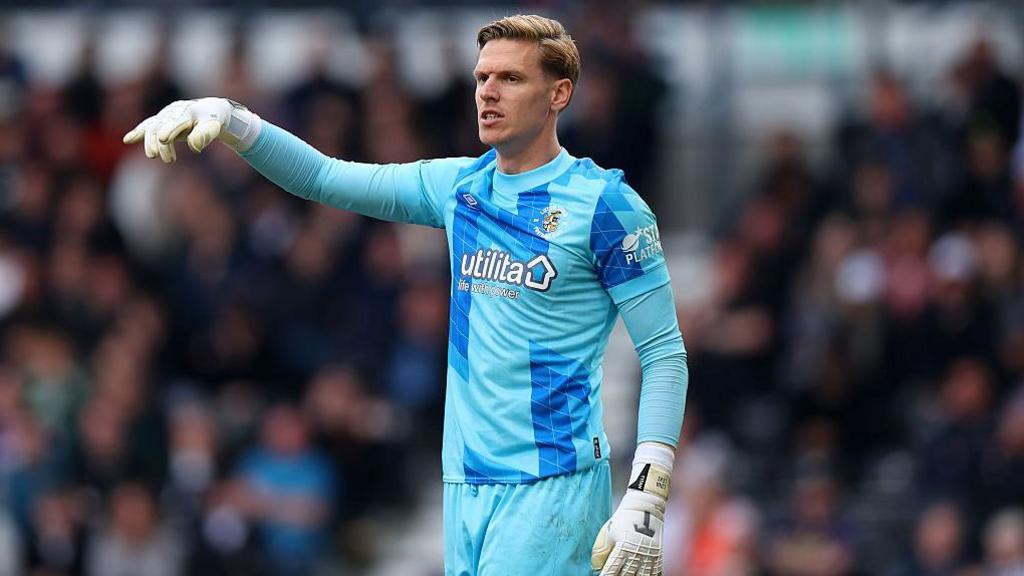 Thomas Kaminski, dressed in Luton Town's patterned turquoise and blue goalkeeper kit, gesturing to a team-mate who is out of picture