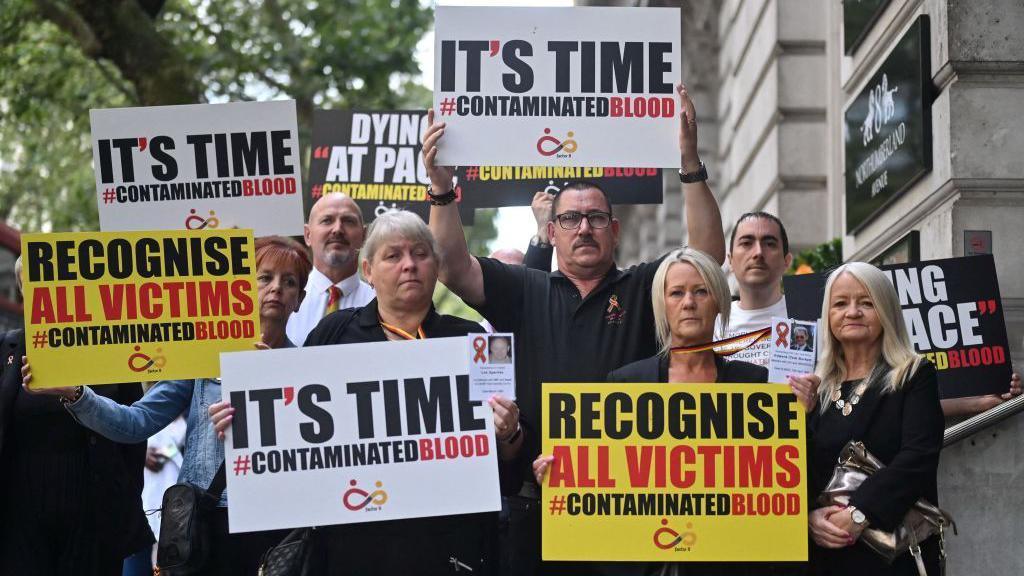 Demonstrators outside the public inquiry in 2023 hold placards related to the infected blood scandal reading: "It's time" and "recognise all victims". 