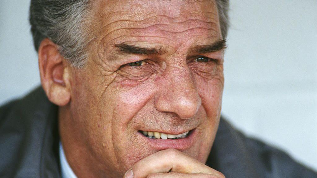 John Lyall looks on from the bench during a match in 1993. He rests his head in his hand. He wears a grey suit with a white shirt and tie. He has short grey hair.