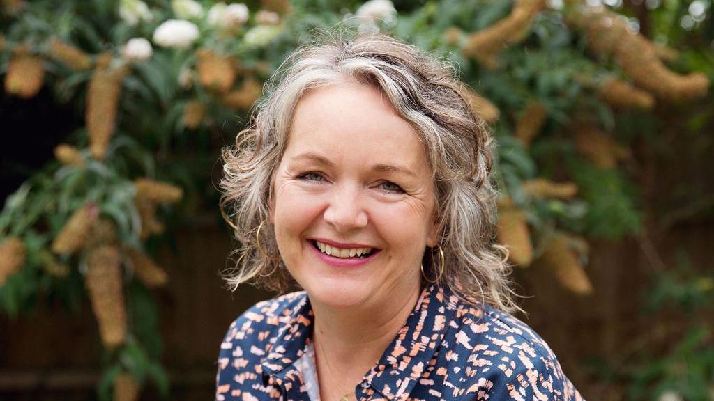 A smiling Alex Allan looks directly at the camera as she is photographed outside before a plant. She has grey hair and is wearing hoop earrings and a multi-coloured shirt.