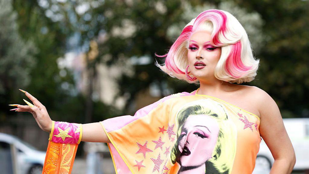 Elle Vosque is wearing a blonde wig with pink highlights. She is wearing an orange off the shoulder dress decorated with pink stars and a picture of Marilyn Monroe. 