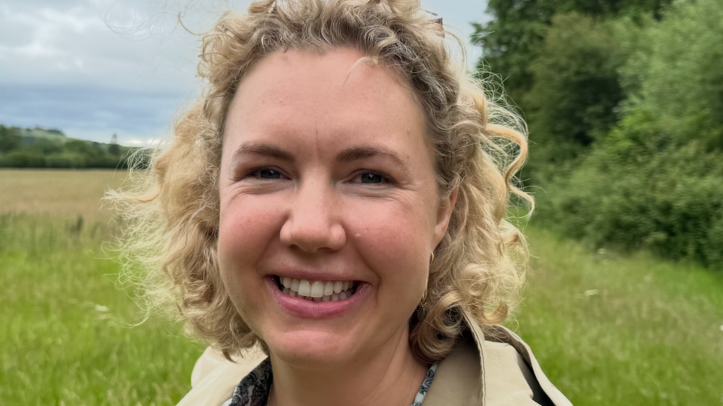 A woman with curly blonde hair is smiling. She is standing in a field covered in wildflowers