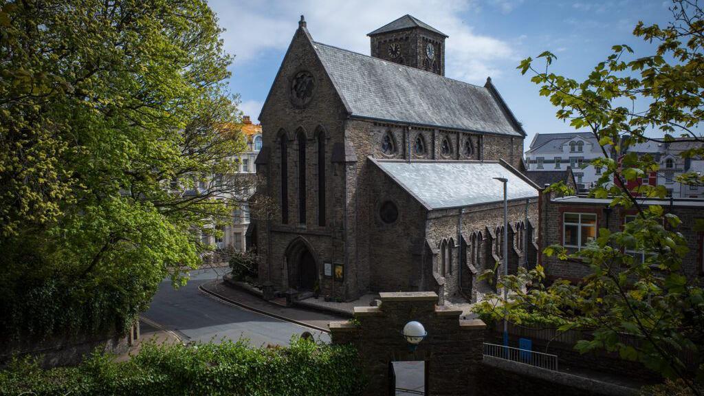 The exterior of St Thomas' Church, which is a stone built church with a steep roof and a clock tower a the rear. There are trees in the fore and a road running past it with blue sky overhead.