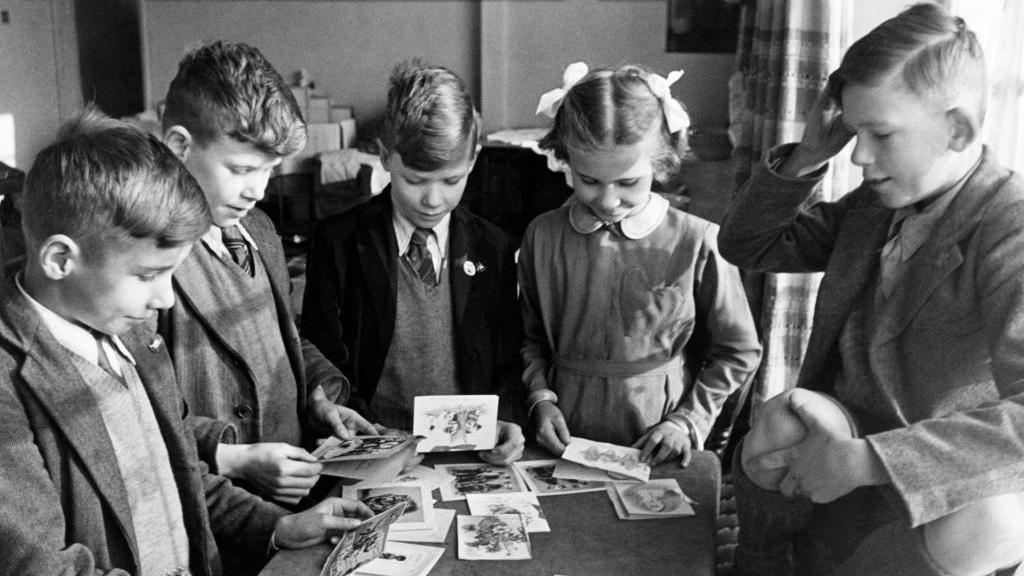 A black and white photo shows four children - three boys and a girl - looking at birthday cards in 1945