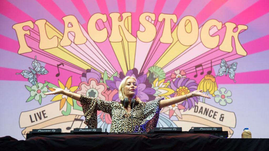 Denise Van Outen, in front of a Flackstock banner, holds her hands out wide as she performs a DJ set at Flackstock in July 2024