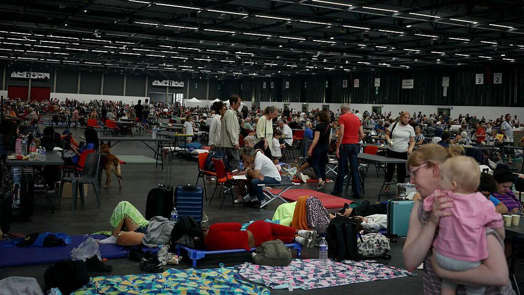 A number of people shelter at an exhibition centre - some people sleep on mats on the floor, while a woman carries a baby on the bottom right of the photo