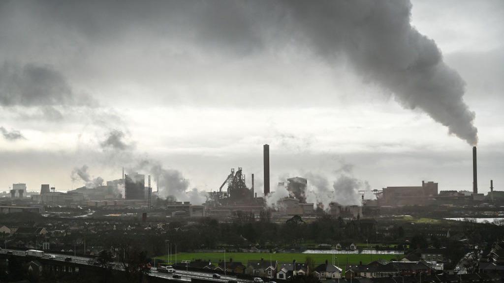 The steelworks at Port Talbot before the blast furnace was closed. Steam and smoke rise from tall chimneys and multiple points across the vast complex. The town and the M4 motorway can be seen in the foreground.