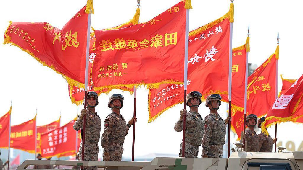 Soldiers in combat uniform stand in the backs of trucks holding large red flags