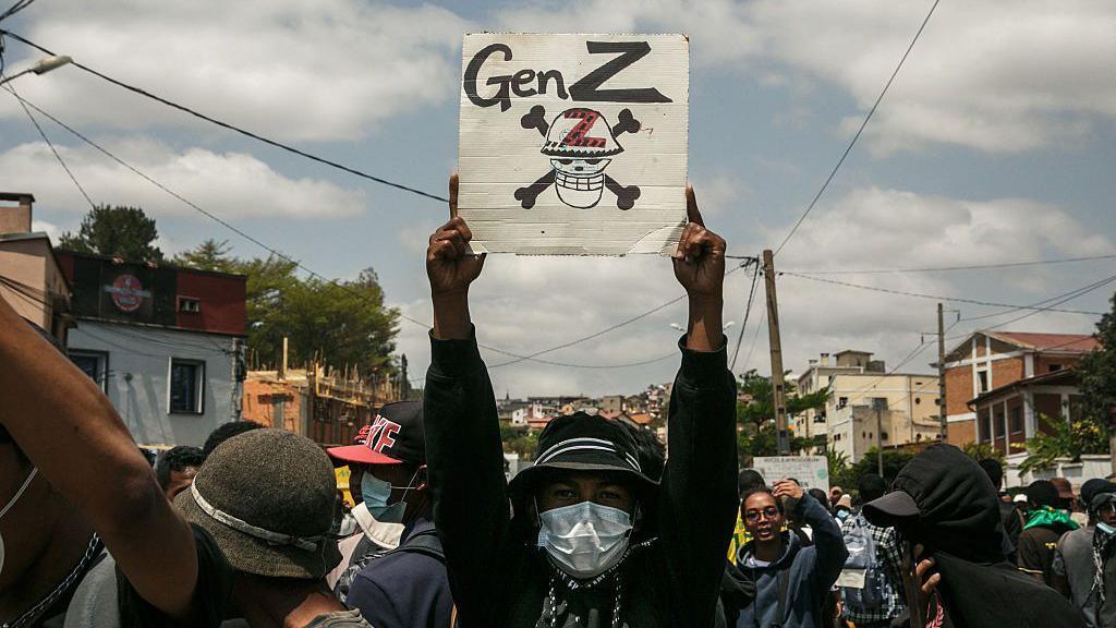 A protester with their face covered by a mask holds up a poster that says 'Gen Z'