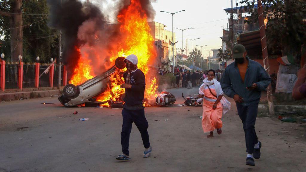 People run past burning vehicles of India's Bharatiya Janata Party (BJP) MLA during a protest to condemn the alleged killing of women and children in Imphal, capital of India's violence-hit northeastern state of Manipur on November 16, 2024. An Indian state riven by months of ethnic tensions imposed an internet shutdown and curfew November 16 after angry protests over the recovery of six bodies of people believed to have been kidnapped by insurgents. (Photo by AFP) (Photo by -/AFP via Getty Images)