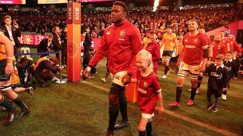 Maro Itoje of the British & Irish Lions, in a red jacket, leads his side on to the pitch before the second test match with Australia at the Melbourne Cricket Ground. He is holding hands with Kate's eldest son, who has his hair tied back in a ponytail.