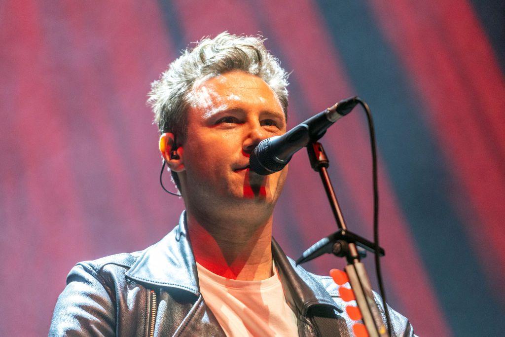 A close-up image of Callum Beattie, in a leather biker jacket and white T-shirt, singing into a microphone during a performance.
