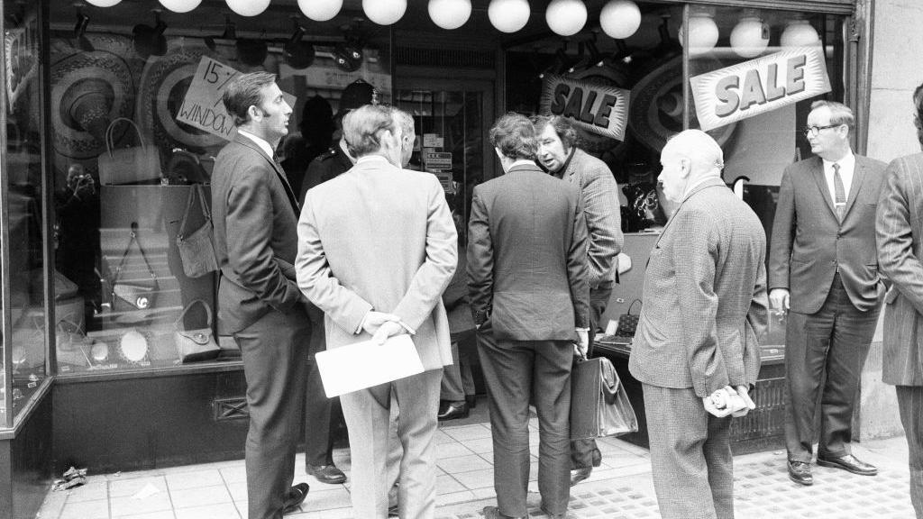 Detectives in suits gather outside SAC, a leather goods shop on Baker Street. They are all looking at each other in a cross manner