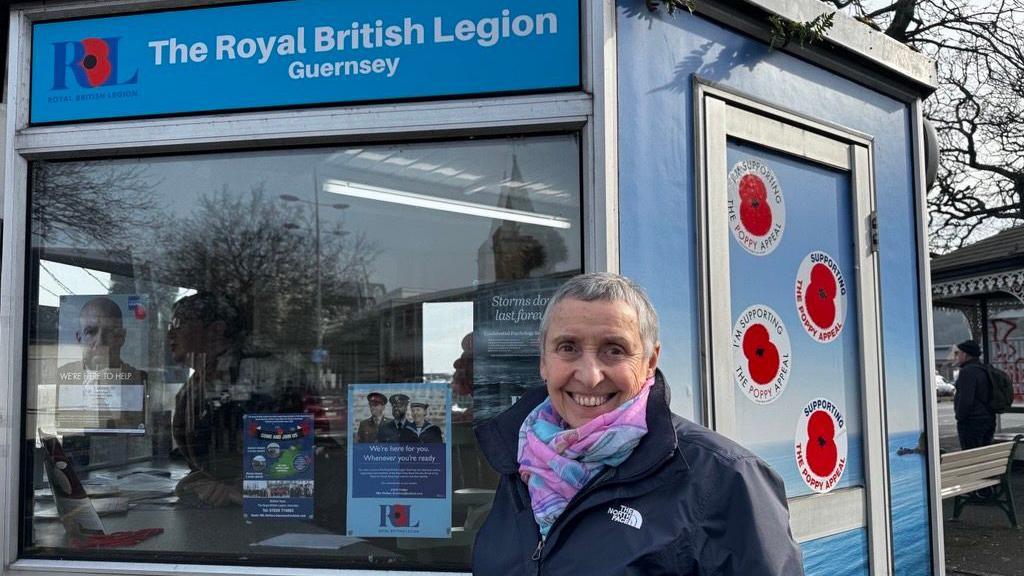 A smiling woman in a blue jacket in front of a hexagonal kiosk, which is decorated with poppy stickers and a Royal British Legion Guernsey sign.