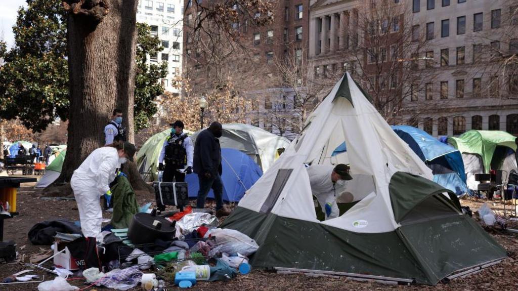 A tent encampment being removed from downtown Washington DC in 2023. Workers in hazmat suits wear face masks. Rubbish is seen strewn outside of one white and green tent. There are large trees and buildings visible outside of the edges of the public park.