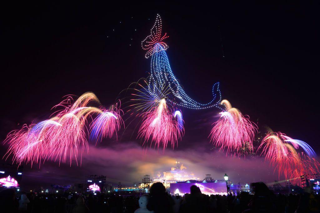 Drones depict an image of a whale and a butterfly against the night sky. They can be seen above seven burst of pink fireworks. A crowd of people are at the bottom of the picture, watching the display. 