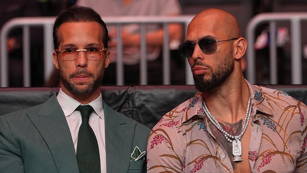 Tristan Tate, in rimless glasses and a grey suit and tie, sits next to Andrew Tate, in a pink open necked shirt, beaded silver chains and dark sunglasses. Both have short facial hair.