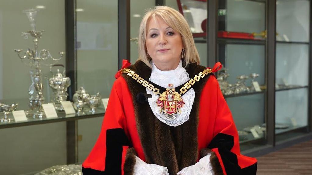 A woman with blonde hair wearing mayoral robes and chain. She is standing in front of cabinets containing the city silver.