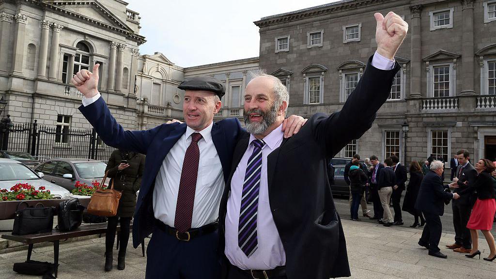 Michael Healy Rae (left) with his brother Danny Healy-Rae (right). Both men are wearing suits and have an arm held in the air. Michael is wearing a hat. 