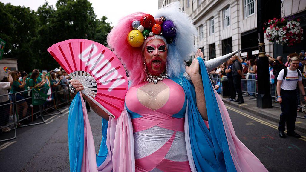 A man with a beard and make up in a pink, white and blue dress with large pieces of artificial fruit on his head. He is carrying a large pink hand fan with the word 'Daddy' written on it in white capital letters. 