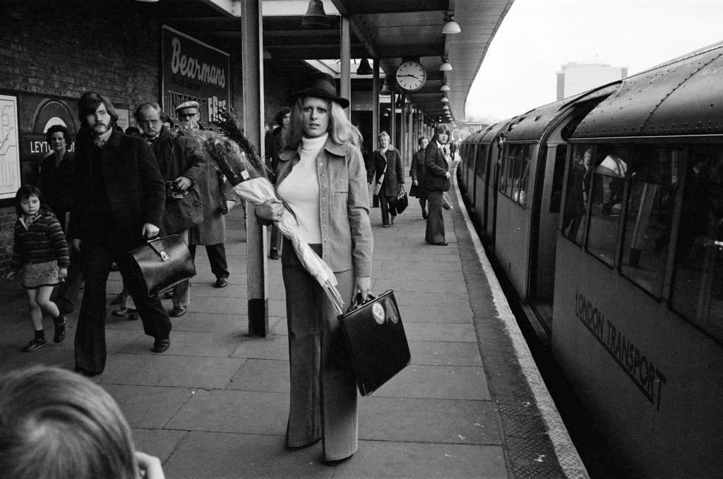 A tall, slim blonde woman wearing a hat and carrying a bunch of flowers in one arm and a flat portfolio case in the other hand. She is at an Underground station and a train can be seen on the right and fellow travellers are on the platform around her.