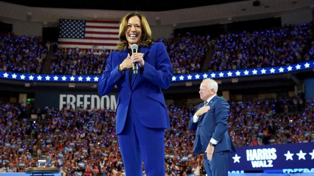 Harris holds a hand to her heart, grinning as she stands on stage at a rally