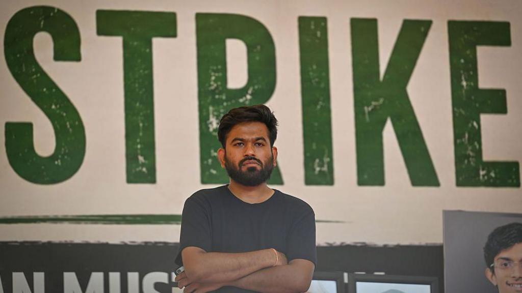 CJP founder Abhijeet Dipke wearing a black T-shirt with his arms crossed. Behind him is a large poster where the word 'STRIKE' can be seen
