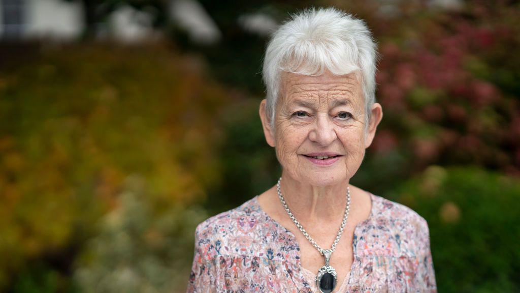 Jacqueline wilson standing outside smiling wearing a pink floral blouse and big silver