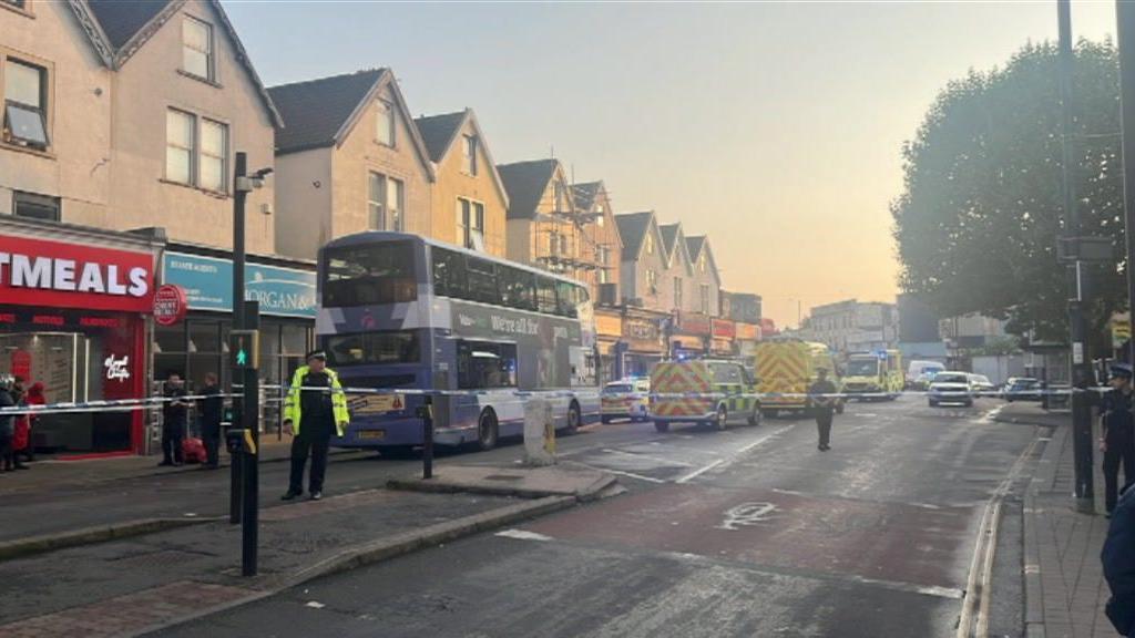 Police scene on a busy road, with officers seen standing behind police tape with ambulances, police cars and vans