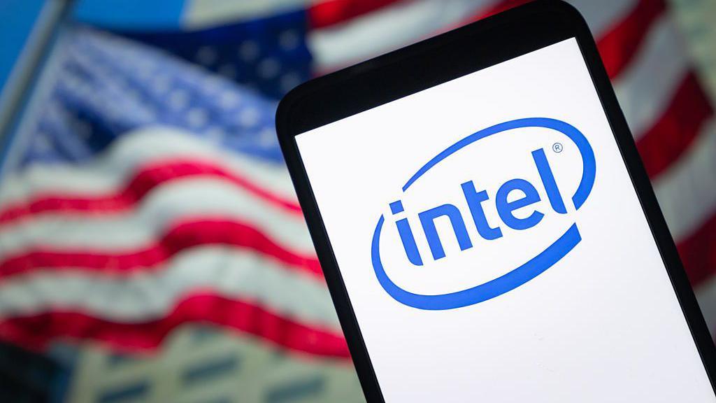 A mobile device displaying the Intel logo appears in front of a blurry US flag