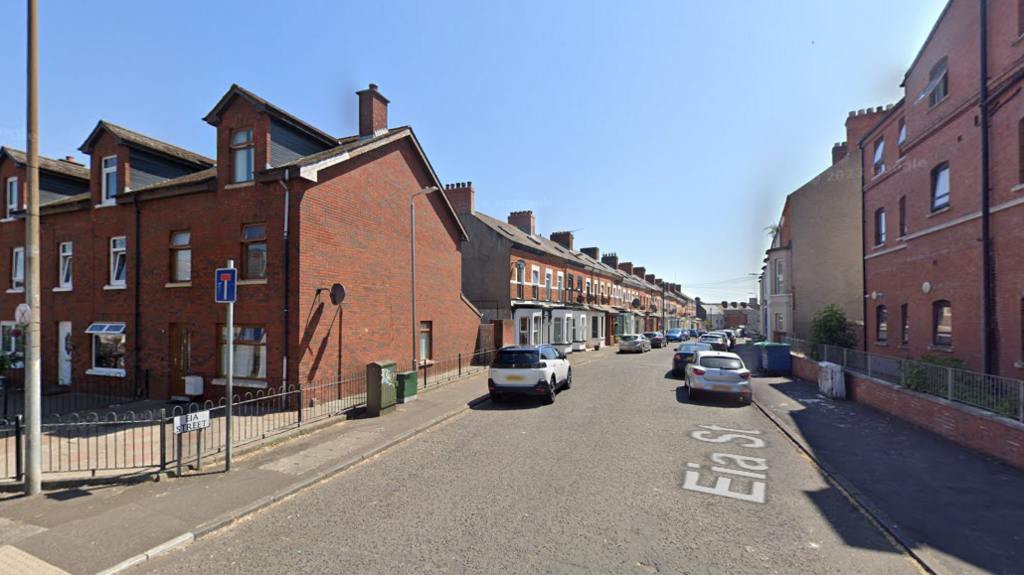 A Google Maps street view of Eia Street in north Belfast. There are a number of cars parked on either side of the street. The street shows a number of red brick, terrace houses with red brick buildings on either side of the street's opening.