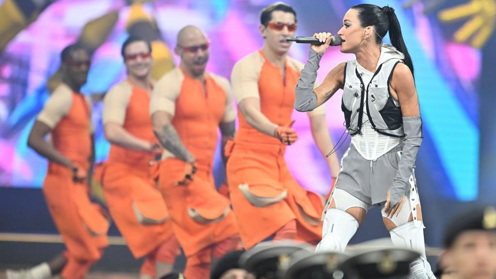 Katy Perry sings into a microphone wearing a futuristic-looking white outfit while backing singers dressed in orange are lined up behind her, in front of a line of military personnel