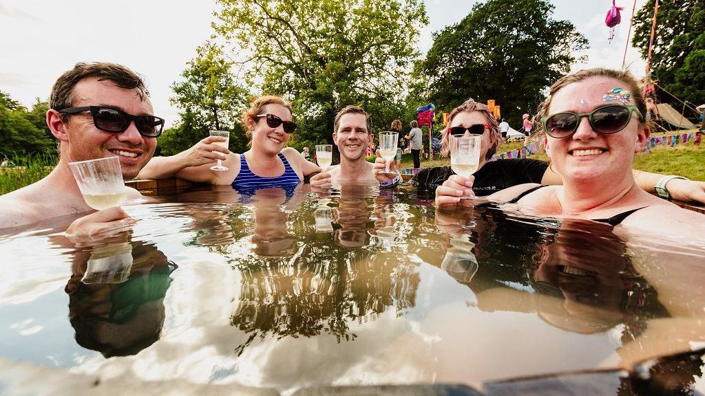 Two men and three women sit in a hot tub in a field with all of them holding glasses of liquid