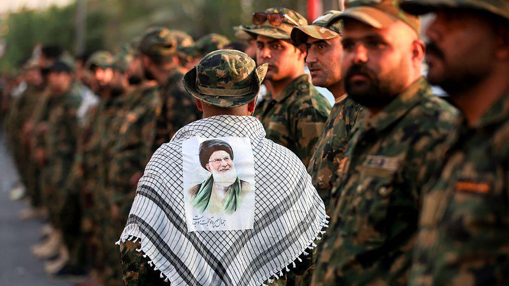 A member of Iraq's Popular Mobilisation Forces (PMF) wears a keffiyeh scarf with a portrait of Iran's late Supreme Leader Ali Khamenei stuck to it, at the shrine of Imam Hussein on the eve of his funeral ceremonies in the holy Iraqi city of Karbala (7 July 2026)