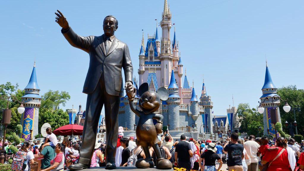 A statue of Walt Disney holding Mickey Mouse's hand in Walt Disney World, Florida, in front of the princess castle. It is a bright sunny day.