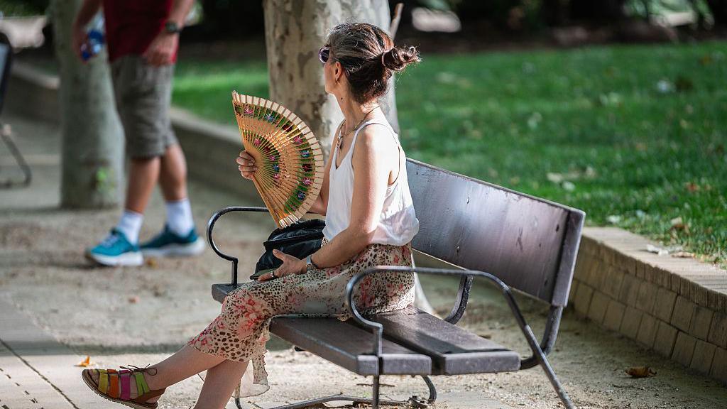 A woman is seen fanning herself while sitting on a park bench during a day of high temperatures resulting from a heatwave
