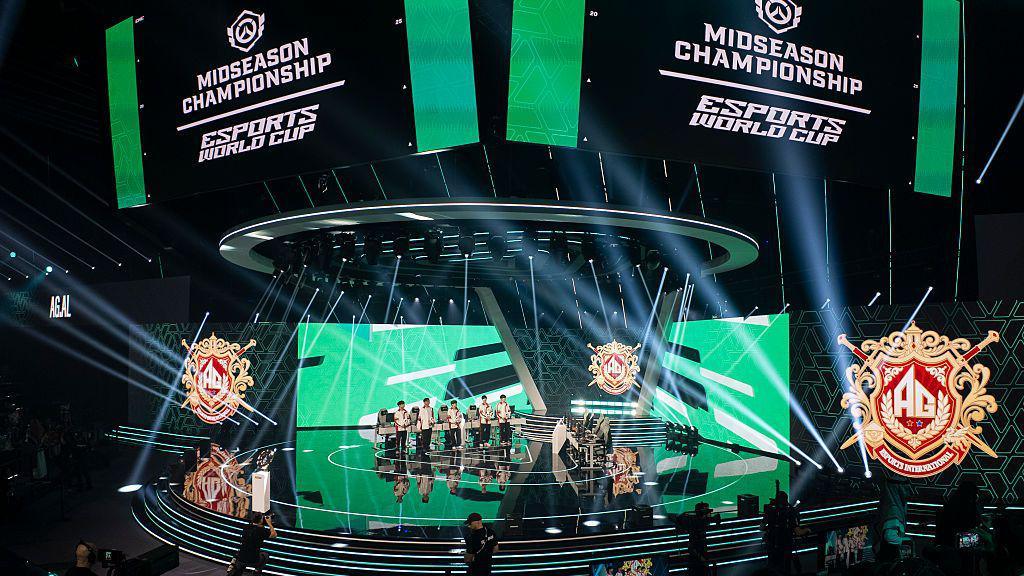 A stage with lights and screens with five esports players standing next to their computer terminals. They all wear white sports tracksuits.