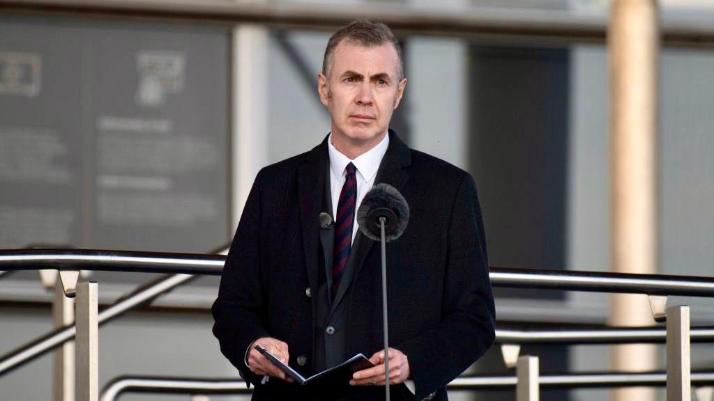 Adam Price in his mid 50s, wearing a black overcoat appearing in front of a microphone to give a speech outside the Senedd.