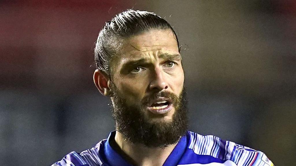 Andy Carroll during a game. He is wearing a blue and white-striped football kit. He has his black hair pulled back into a bun and has a bushy dark beard. He is sweaty during a football match.