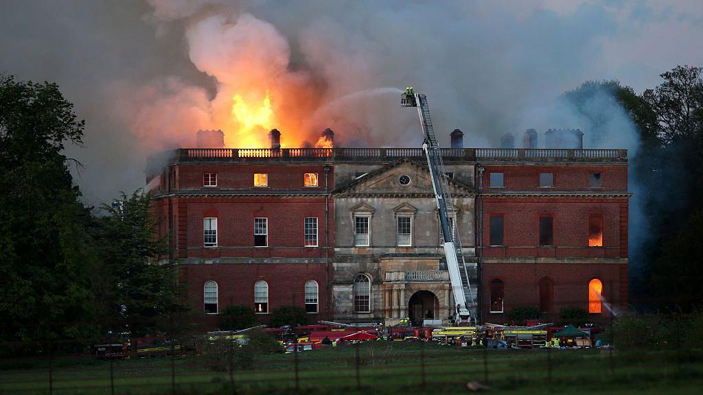 Fire engines surround the house which is seen from the distance with large flames coming out of its left hand roof and also seen through lower windows on the right side of the building.