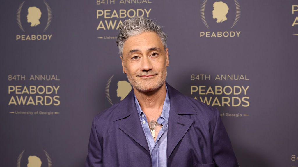 Taika Waititi in a purple jacket smiling at the camera.