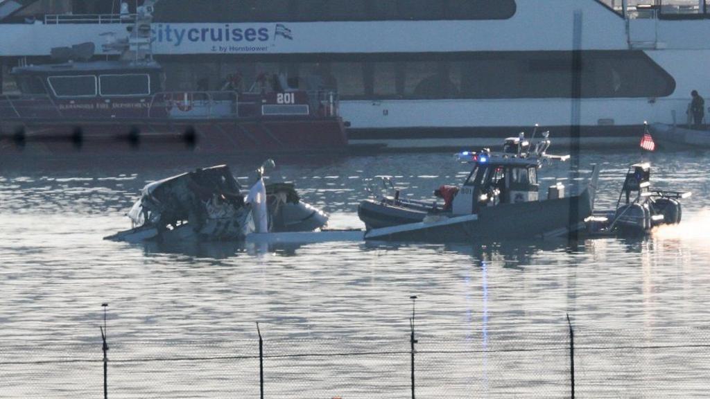 Two  boats next to the wing of the plane in the water, as seen from the shore. A City Cruises vessel is in the background