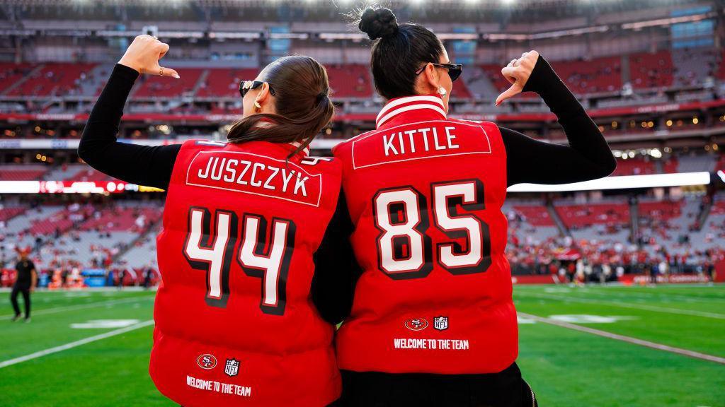 Two women in an American football stadium. They are wearing bright red puffer jackets. On the left, the woman's jacket has the number 44 on it, and says Juszczyk. On the right, the woman's jacket has the number 85, and says Kittle.