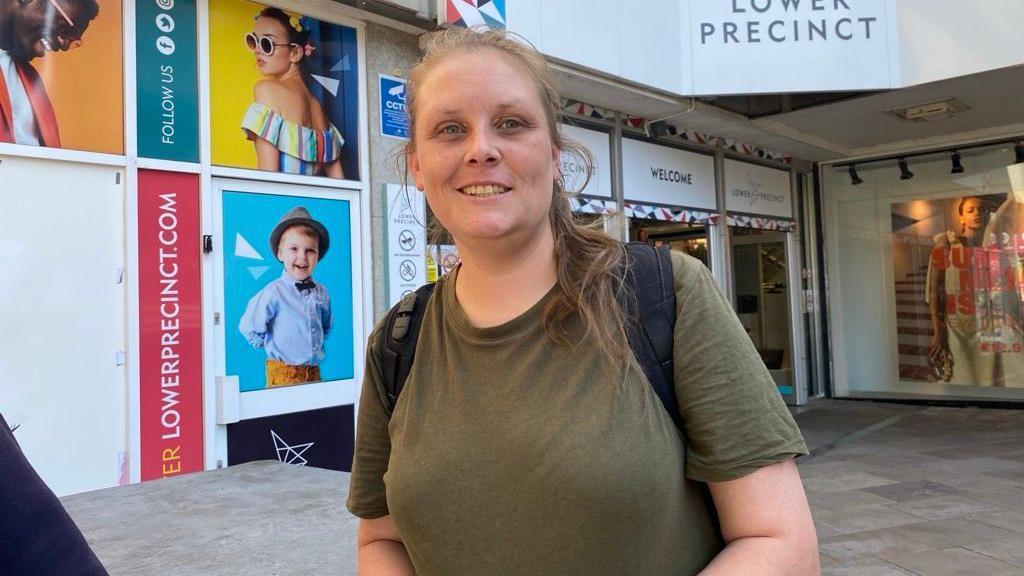A woman wearing a green T-shirt and black rucksack looks towards the camera. A building and shop signs with the text Lower Precinct, is in the background behind her.